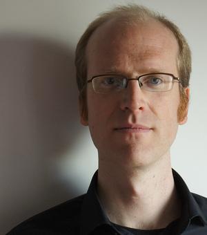 Koen Vermeir looking directly at the camera. They are wearing a dark shirt and are standing against a light wall with glasses on.