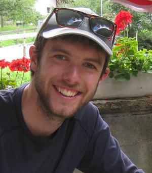 Image of Tim Middleton smiling, wearing a hat with small boxed red flowers behind him