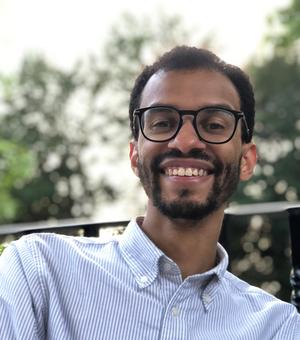 Michael joseph, smiling broadly at the camera. he is wearing a white and blue striped shirt and dark glasses