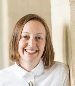 Ruth Scobie smiles at the camera against a light stone background.