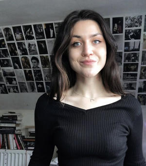 woman with long brown hair wearing a black top in front of wall with pictures behind