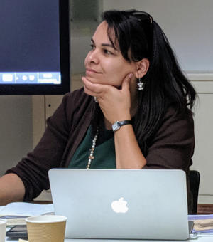 rosa andujar looking left wearing dark green, hand to chin,an apple laptop sits in front of her. 