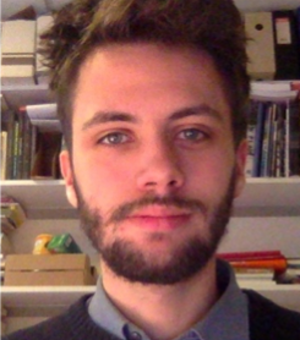 Headshot of a man wearing a shirt and jumper stood in front of a bookcase