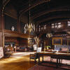 A photograph showing the large, wood-panelled interior of the Great Parlour at Wightwick Manor, Wolverhampton, filled with Arts and Crafts furniture and decorative objects. The wooden ceiling features decorative painting and two large brass chandeliers.
