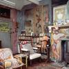 A cluttered corner of an indoor art studio - walls painted in various block colours, two chairs with multicoloured cushions, paintings and sculptures covering the walls. A bookshelf in the background and an unlit fireplace with a tiled and painted facade 