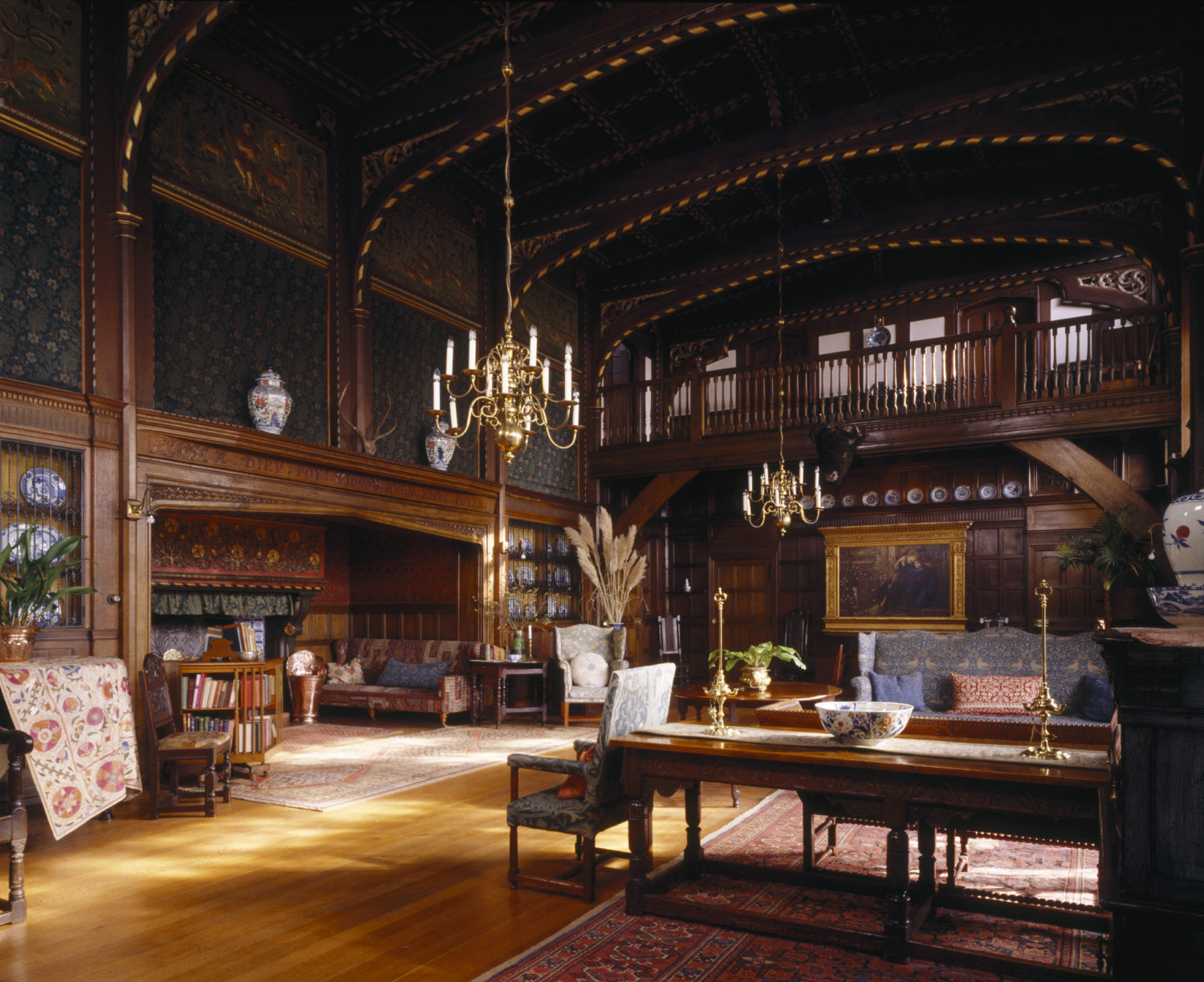 A photograph showing the large, wood-panelled interior of the Great Parlour at Wightwick Manor, Wolverhampton, filled with Arts and Crafts furniture and decorative objects. The wooden ceiling features decorative painting and two large brass chandeliers.