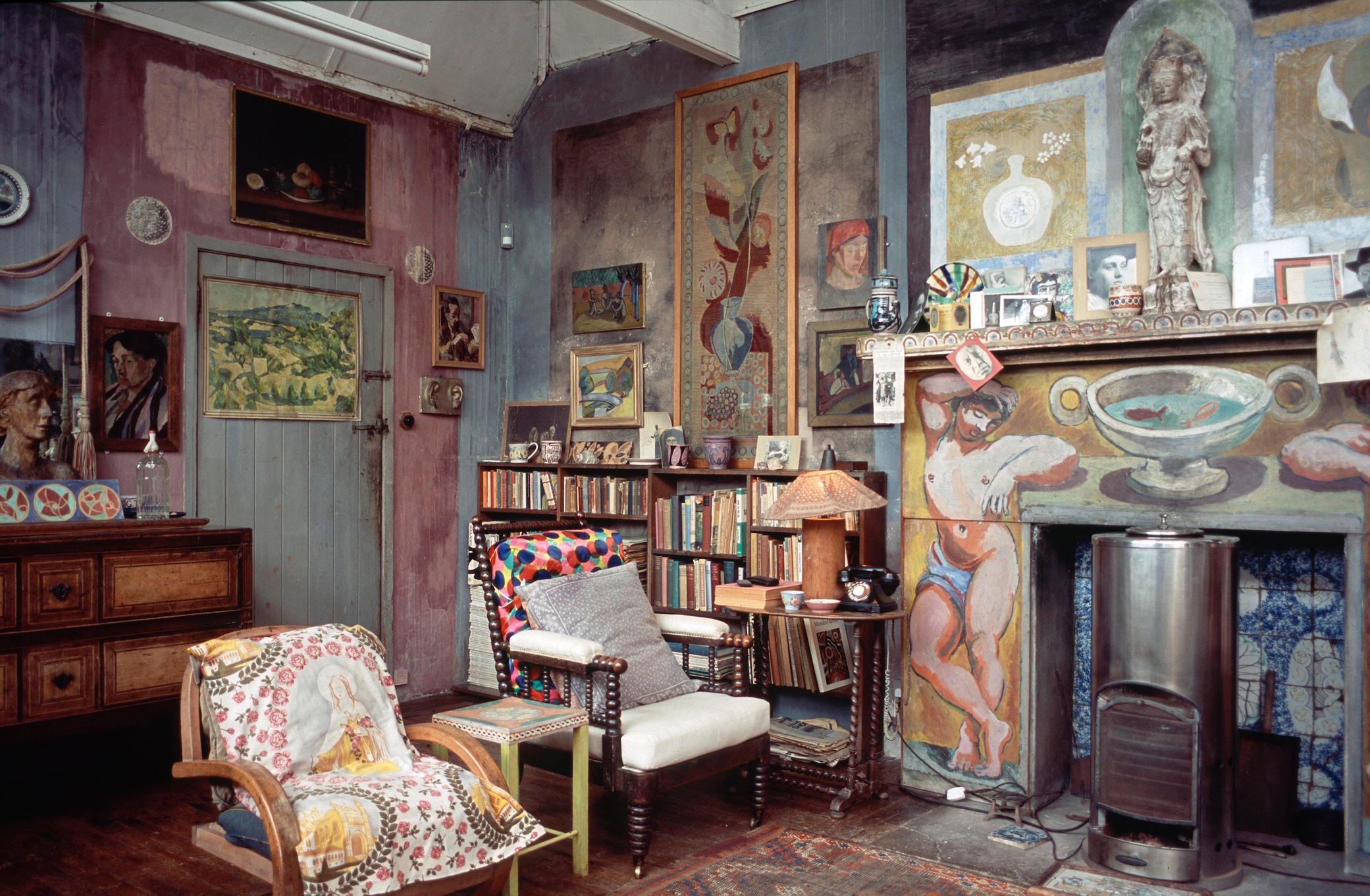 A cluttered corner of an indoor art studio - walls painted in various block colours, two chairs with multicoloured cushions, paintings and sculptures covering the walls. A bookshelf in the background and an unlit fireplace with a tiled and painted facade 