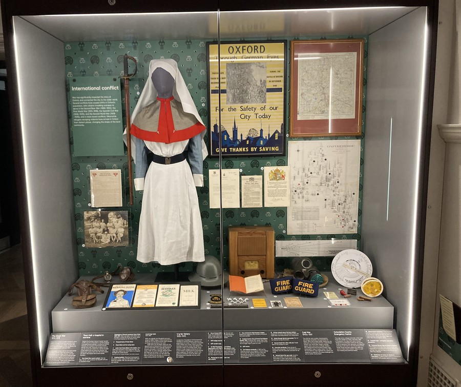 A grey square museum display case is filled with dozens of objects relating to various conflicts. 