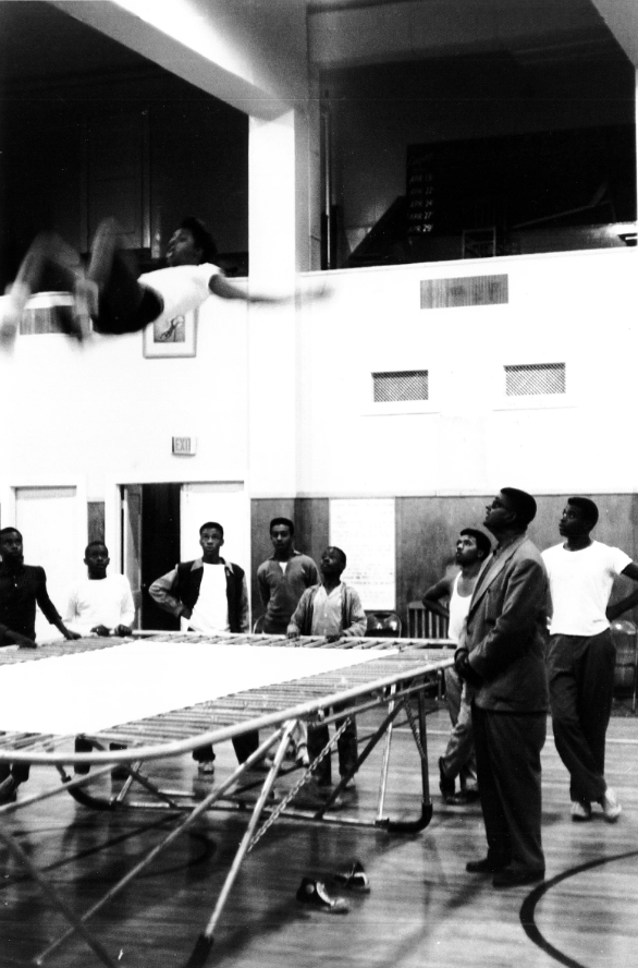 A person mid-bounce on a trampoline, others stand looking up at him.