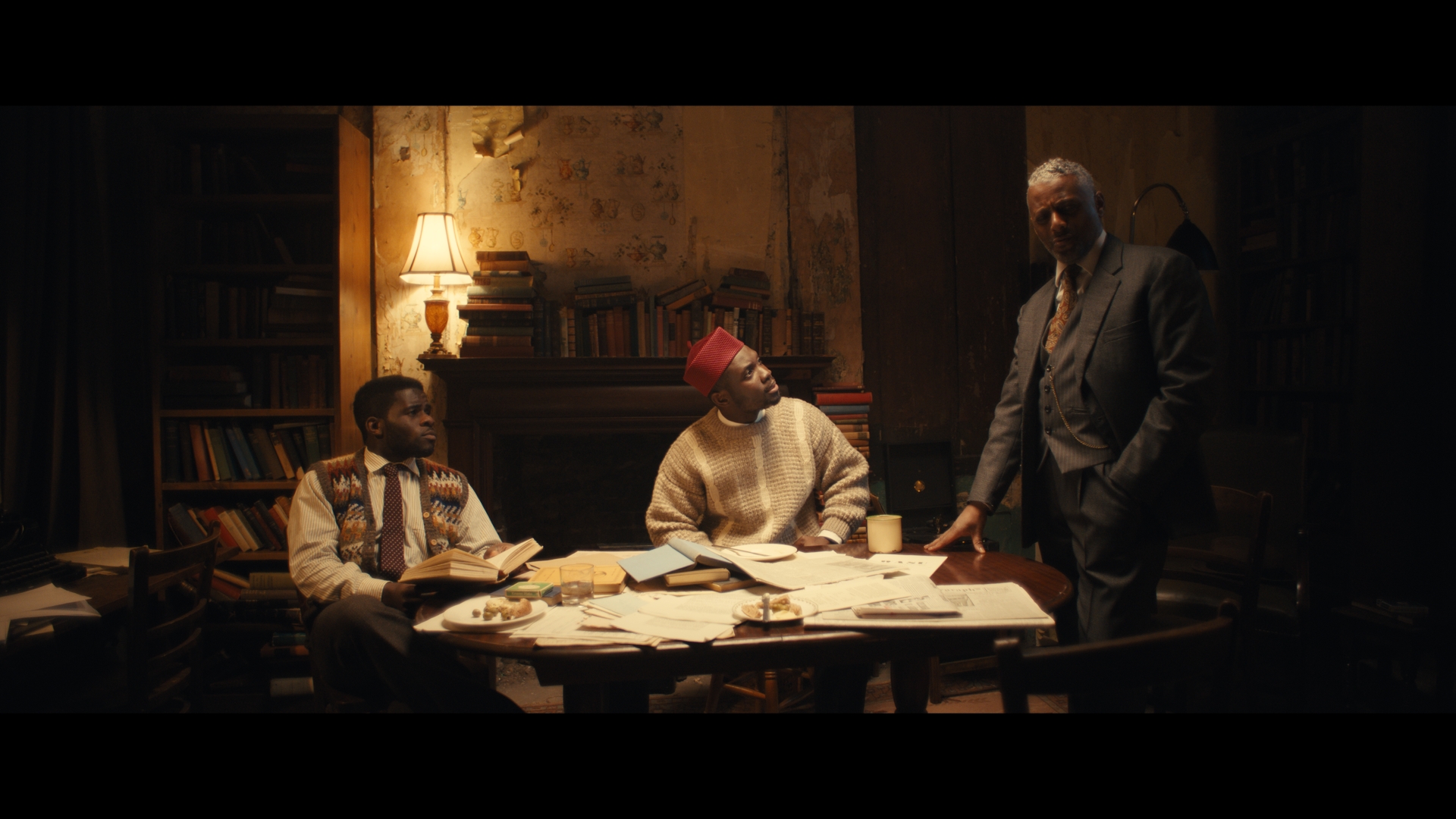 Three men wearing period clothes are sitting in a room at a table. Two of them are sitting and one of them to the extreme right is standing.